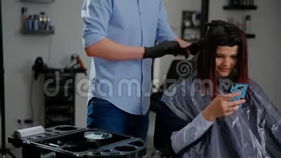 美容发型和人的概念幸福的年轻女性有智能手机和理发师在美容院做发型视频的预览图