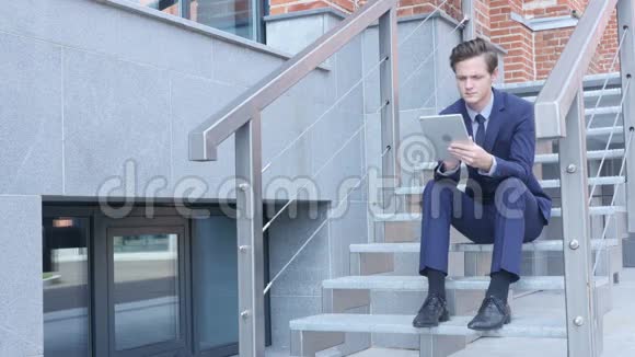 年轻商人使用平板电脑浏览坐在办公室外的楼梯上视频的预览图