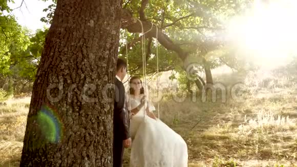 快乐的新郎在夏天公园里穿着漂亮的白色连衣裙在摇摆新娘上摇摆在夏日森林的橡树枝上荡秋千视频的预览图