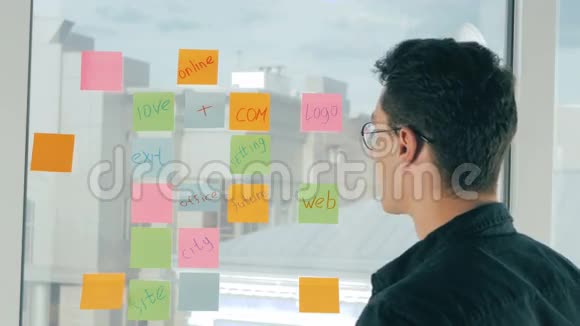 年轻的帅哥在现代办公室的全景窗口上写着粘稠的笔记头脑风暴命名视频的预览图