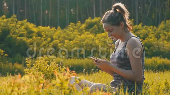 坐在绿色草坪上在日落背景下的风景区使用智能手机的快乐年轻女子视频的预览图