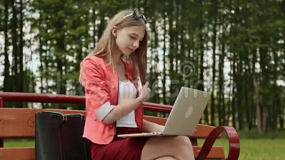美丽的年轻女孩金发放在公园的长凳上在她的笔记本电脑上工作女孩用笔记本电脑打字视频的预览图