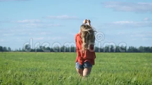 一个美丽的金发年轻浪漫的女人穿着红色衬衫在绿色的田野上慢慢行走的肖像从后面看视频的预览图