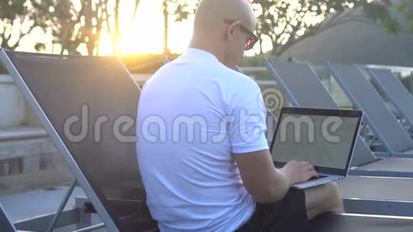 户外照片一个英俊的年轻人坐在屋顶的日光浴床上在笔记本电脑上工作视频的预览图