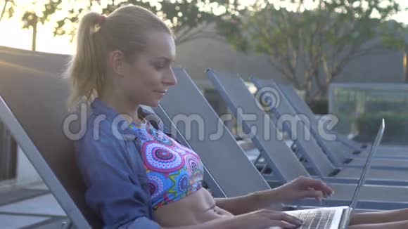 穿着比基尼和牛仔裤衬衫的金发美女坐在屋顶的日光浴床上用笔记本电脑工作视频的预览图