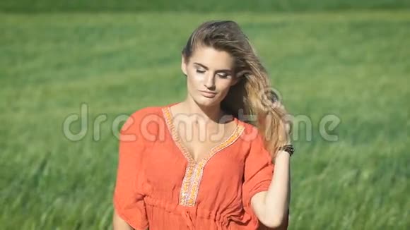 一位美丽的金发年轻浪漫女子穿着红色衬衫在绿茵场上抚摸着她的头发视频的预览图