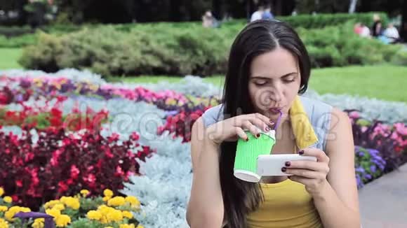 美丽的黑发女人在公园里等着喝咖啡玩智能手机打电话微笑花夏天好天气奶酪视频的预览图