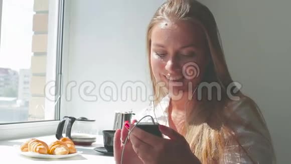 年轻漂亮的女孩在晴天吃早餐听音乐使用智能手机视频的预览图