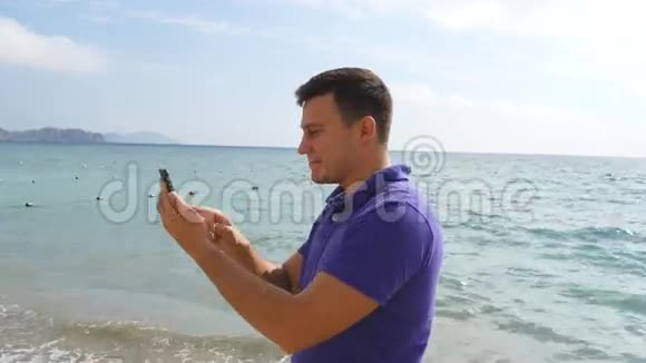 微笑的年轻人在海边用智能手机打视频电话快乐的男人在网上聊天视频视频的预览图