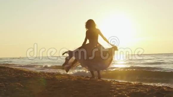 年轻迷人的女孩穿着长长的浅色连衣裙在金色的日落光线照射下在沙滩上旋转视频的预览图