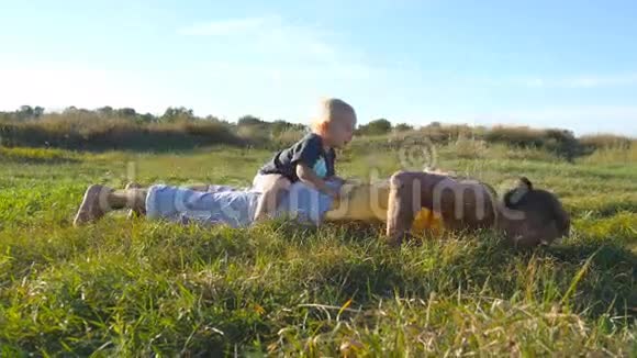 年轻强壮的父亲做俯卧撑他的小儿子在他的背上运动员在草地上做俯卧撑视频的预览图