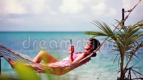 年轻可爱的女孩戴着太阳镜躺在户外秋千床上用手机享受日光浴1920x1080视频的预览图