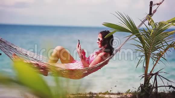 快乐的海滩女人戴着太阳镜在智能手机上发短信躺在吊床上放松热带度假视频的预览图