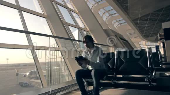 身着休闲装的英俊微笑男子的肖像坐在机场候机楼大厅时他的平板电脑视频的预览图