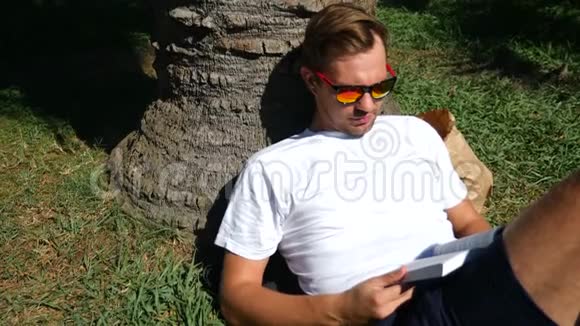 帅哥坐在公园的草地上树下有一棵树看书4k慢动作视频的预览图
