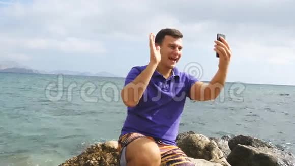 微笑的年轻人在海边用智能手机打视频电话快乐的男人在网上聊天视频视频的预览图
