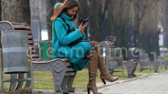 穿着蓝色外套的年轻女孩坐在长凳上用智能手机视频的预览图