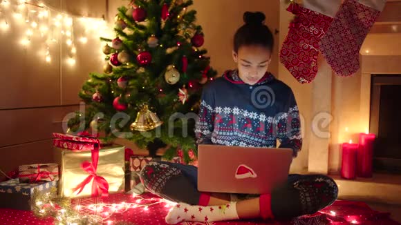 年轻的黑人妇女使用笔记本电脑上网聊天坐在装饰好的圣诞树旁视频的预览图