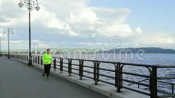 穿着运动布的白种人黑发女人在河边的河堤上奔跑视频的预览图