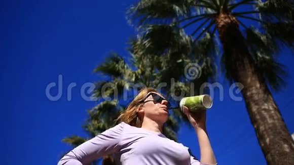 女孩正在街上喝一杯饮料从一个一次性的杯子靠在蓝天和棕榈树上基本观点视频的预览图