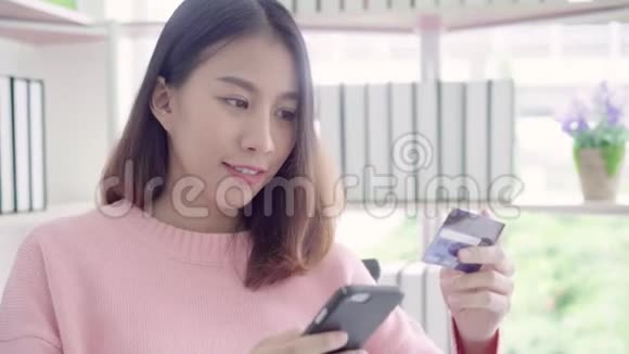 美丽的亚洲女人使用智能手机通过信用卡购买网上购物而穿毛衣坐在客厅的桌子上视频的预览图