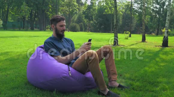 一个穿着蓝色衬衫的年轻人坐在公园的椅子上在电话里得到了一条信息视频的预览图
