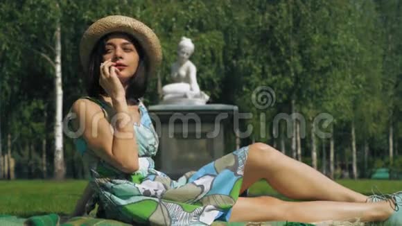 一个戴着帽子的年轻女孩坐在公园草坪上打电话微笑着说话视频的预览图