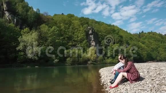 年轻夫妇英俊的年轻人和漂亮的女孩在水中滴石头坐在鹅卵石河岸上视频的预览图