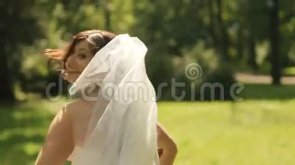 美丽的新娘穿着华丽的白色连衣裙带着长长的面纱在公园的草坪上嬉戏地跑掉了视频的预览图