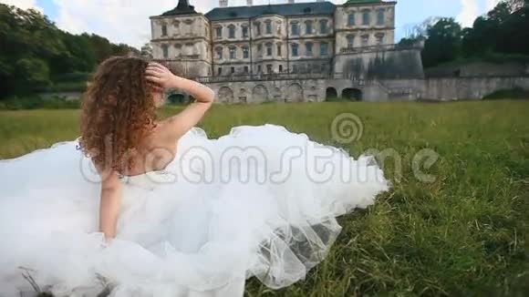 美丽的年轻新娘穿着白色婚纱在绿草如茵的城堡附近看着镜头视频的预览图