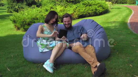 一对年轻夫妇坐在搁脚凳公园的椅子上在平板电脑上观看照片视频的预览图