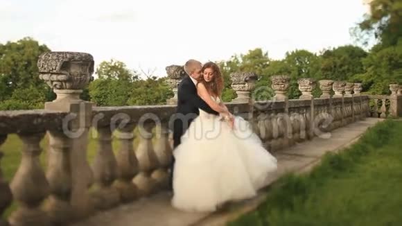 可爱的新郎把他美丽的新娘抱在老石栏杆附近的绿色草坪上亲吻她的脖子视频的预览图