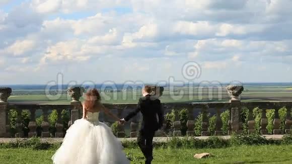可爱的新郎和美丽的新娘走在老石栏杆附近的绿色草坪上大片的蓝天视频的预览图