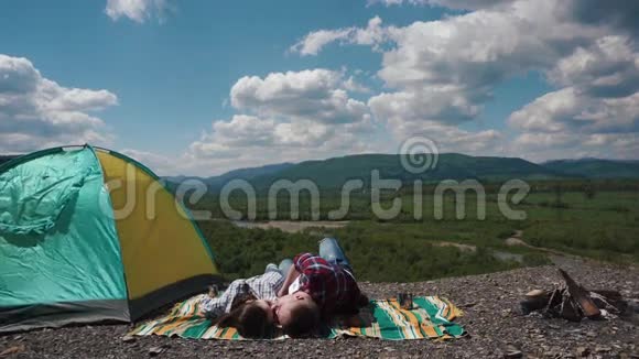 年轻的旅游情侣在蓝底青山和山谷背景下的露营地躺在毯子上接吻视频的预览图