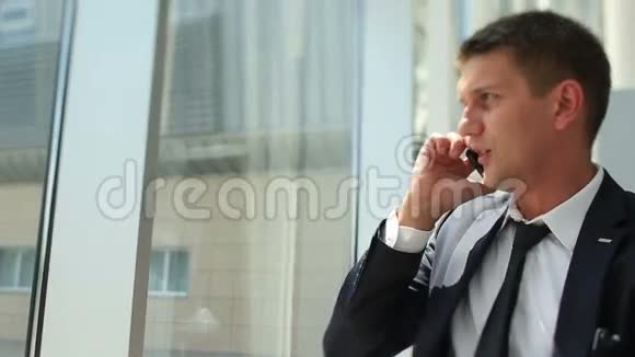 英俊的年轻商人笑着在电话里说话看着现代建筑的窗户视频的预览图