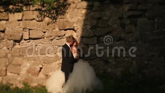 可爱的新郎温柔地抱着他美丽的新娘拥抱她站在古色古香的破墙旁视频的预览图