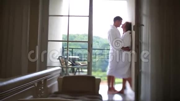 可爱的白种人夫妇穿着浴袍在阳台上拥抱和亲吻视频的预览图