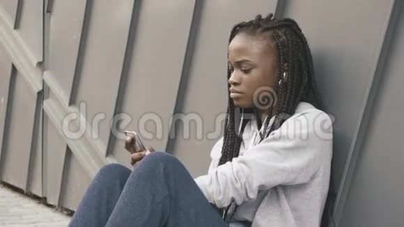一位年轻美丽的非洲女孩坐在灰色纹理的墙边选择音乐倾听的肖像视频的预览图