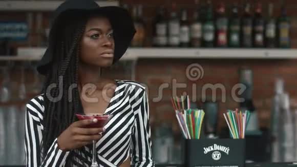 时尚的非裔美国女士戴着黑色帽子和条纹时尚连衣裙在酒吧喝鸡尾酒夏日炎炎视频的预览图
