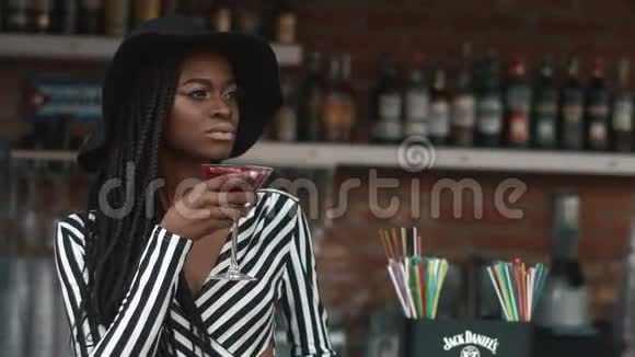 穿着黑色帽子和条纹连衣裙在酒吧里喝鸡尾酒的时尚优雅的非洲裔美国妇女视频的预览图