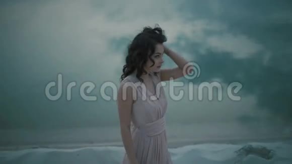 年轻的性感黑发女孩穿着淡粉色连衣裙在沙质海岸散步时玩着面纱摸着她的脸视频的预览图