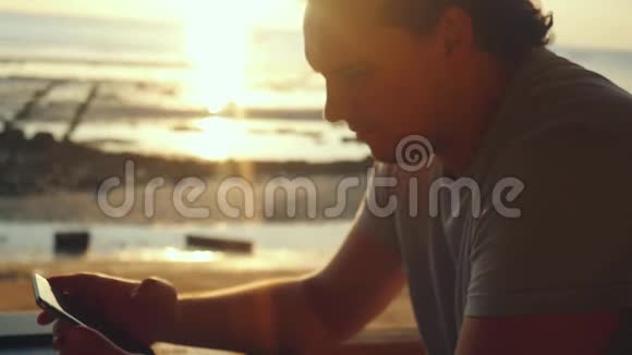 在海滩上的酒吧休息的英俊男子在美丽的日落中缓慢地使用手机1920x1080视频的预览图