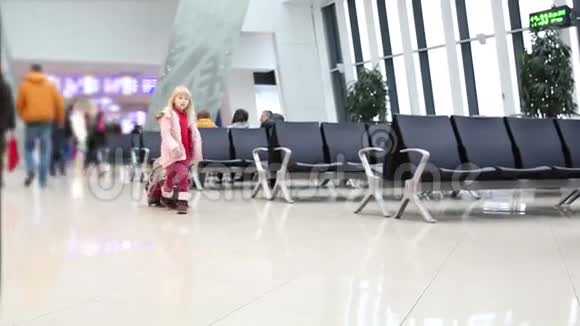 穿着粉红色冬衣的金发小女孩带着红色的手提箱穿过机场的候机大厅视频的预览图