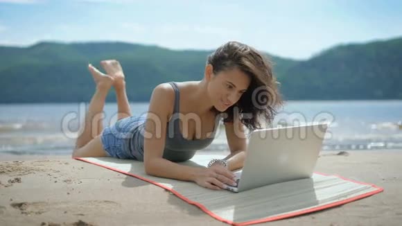 一位美丽的年轻微笑的女士正在用笔记本电脑躺在海滩上的汤米身上视频的预览图