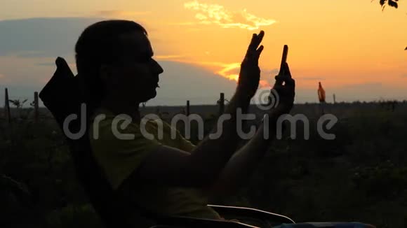 一个男人拿着电话坐在椅子上在日落后橙色天空的背景下视频的预览图
