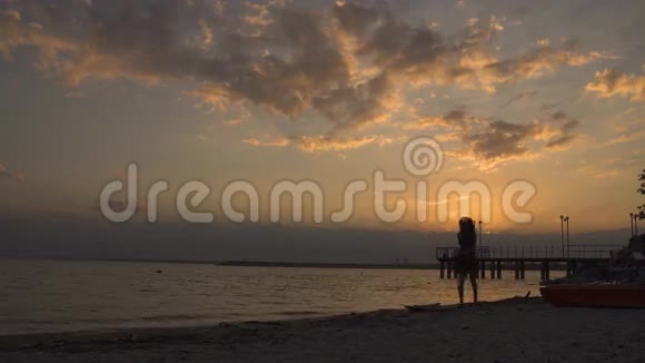 日落时海滩上一个穿着浅色连衣裙的女孩的剪影美丽的少女赤脚走在沙滩上视频的预览图