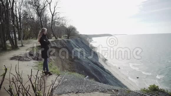 一个年轻的女人从岩石上看着大海等待着什么穿着外套留着长发蓝天视频的预览图
