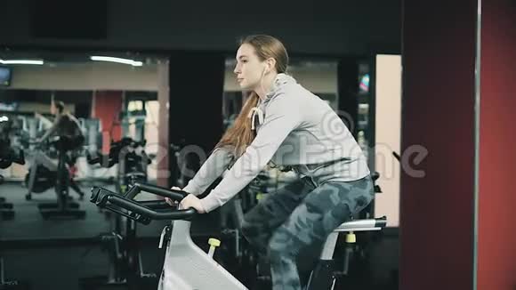 年轻漂亮的女孩在健身房在自行车模拟器上摇动她的腿对着镜头微笑概念热爱运动视频的预览图