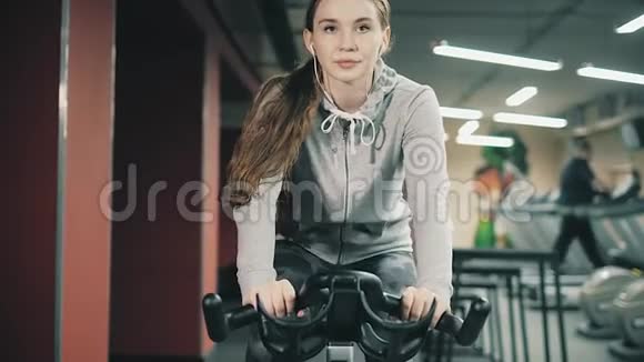 年轻漂亮的女孩在健身房在自行车模拟器上摇动她的腿对着镜头微笑概念热爱运动视频的预览图