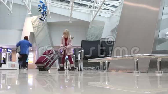 穿着粉红色冬衣的金发小女孩带着红色的手提箱穿过机场的候机大厅视频的预览图
